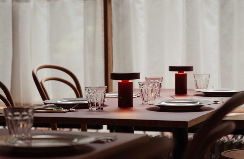 Gedeckter Tisch mit zwei Tischlampen, Fenster mit weißem Vorhang im Hintergrund.